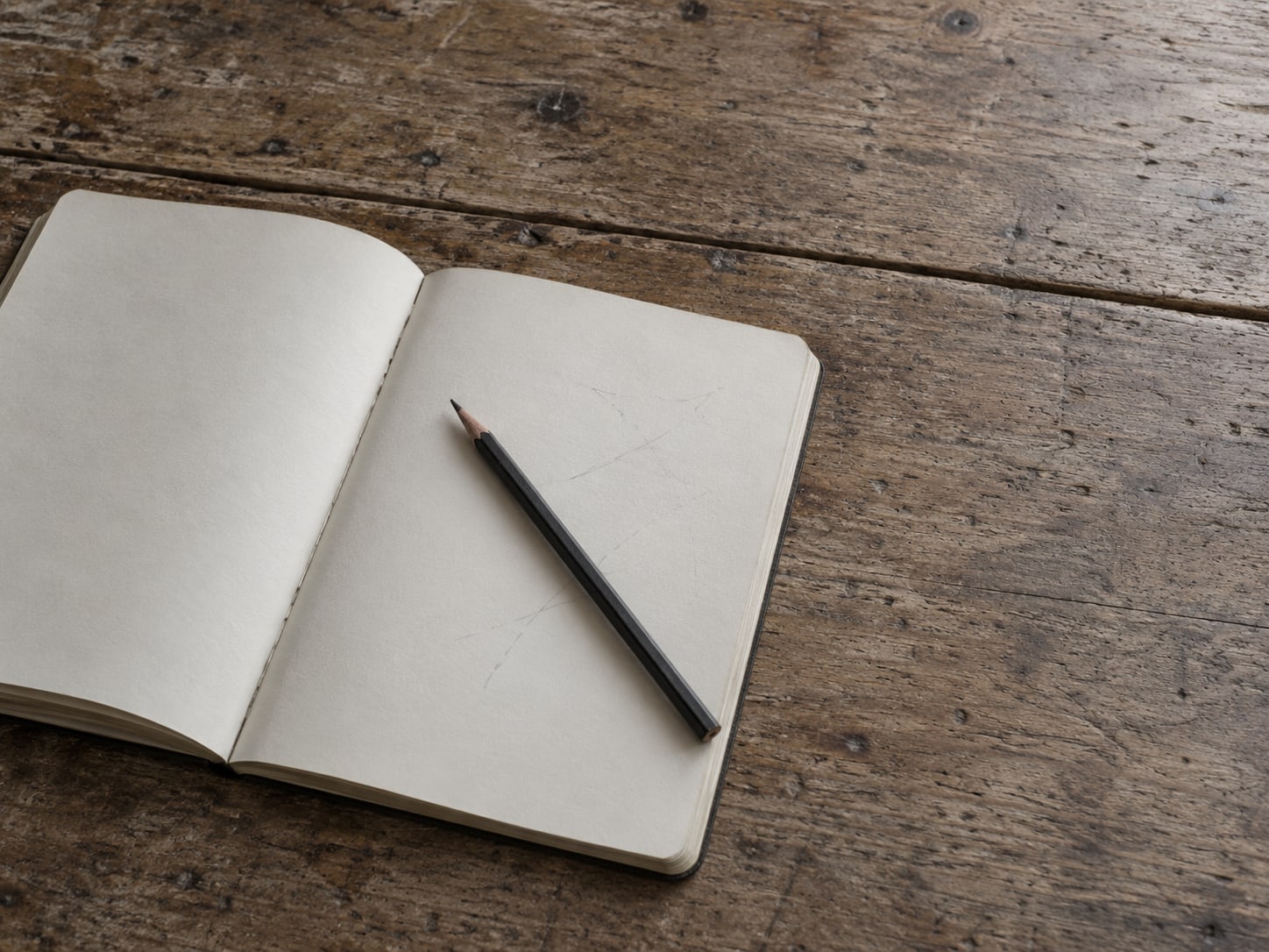 An open blank notebook with a pencil resting across it on a wooden desk, a few faint pencil marks on the page.