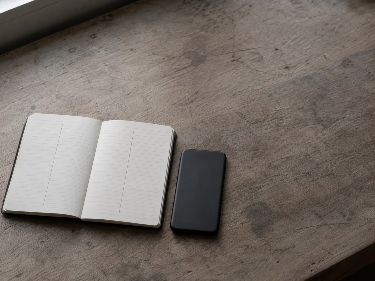 A notebook open to a ruled page with two unreadable pencil columns beside a closed phone lying face down on a desk.