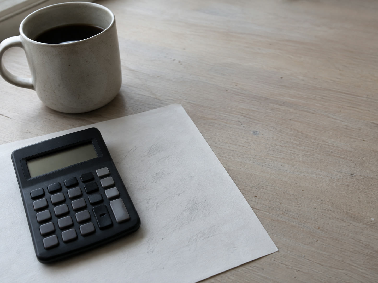 Una pequeña calculadora manual con la pantalla en blanco sobre una sola hoja de papel cubierta de marcas de lápiz tenues e ilegibles junto a una taza de café simple.