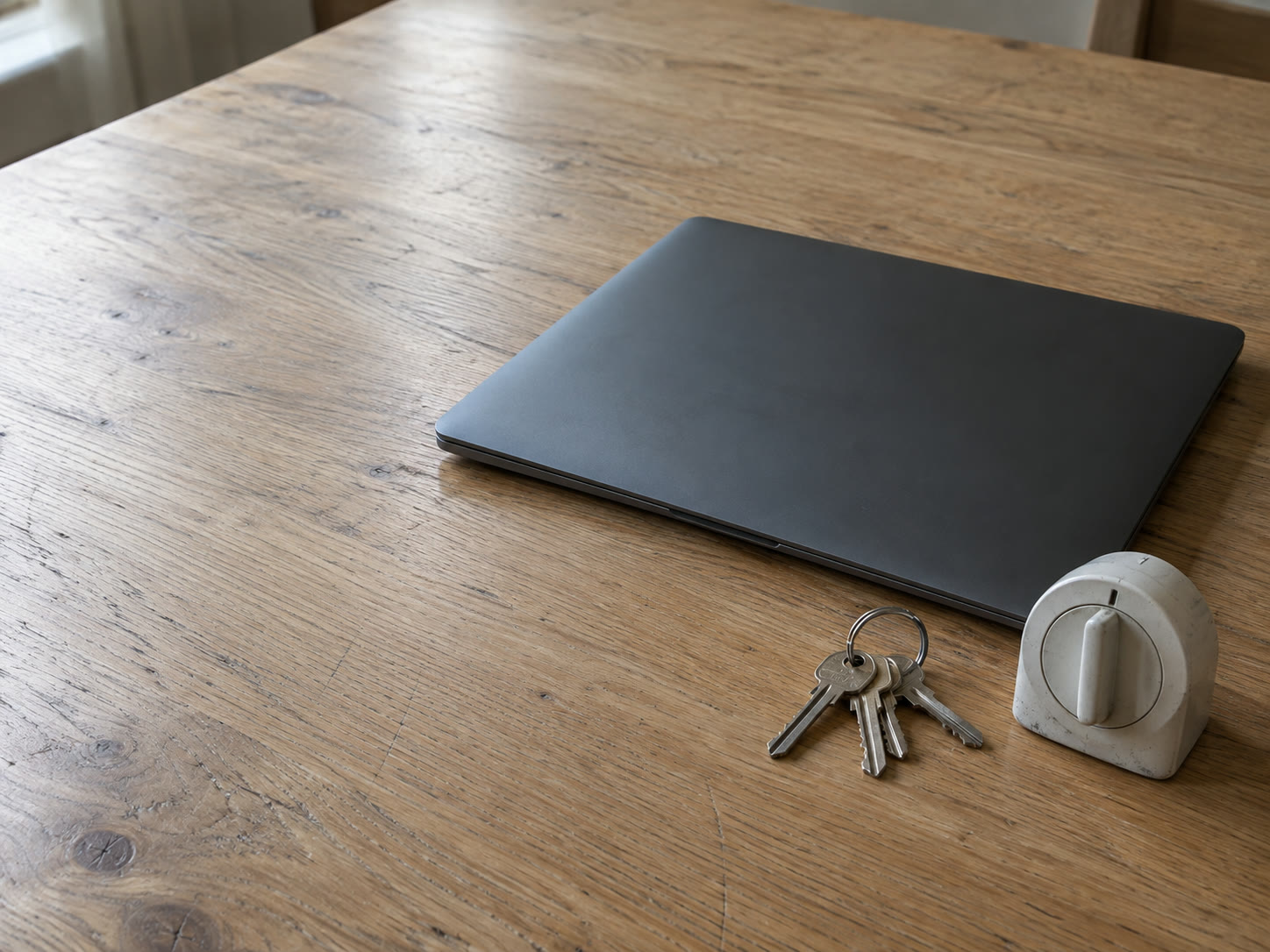 Una laptop cerrada descansando junto a un llavero simple y un pequeño temporizador de cocina sobre una mesa limpia de roble con luz suave de día.