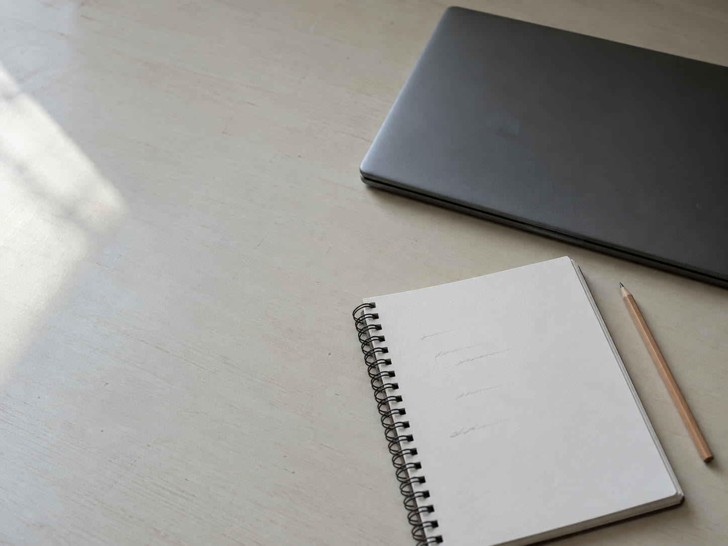 Un cuaderno de espiral abierto en una página de líneas cortas escritas a mano en lápiz demasiado tenues para leer, junto a una laptop cerrada y un lápiz afilado sobre un escritorio pálido.