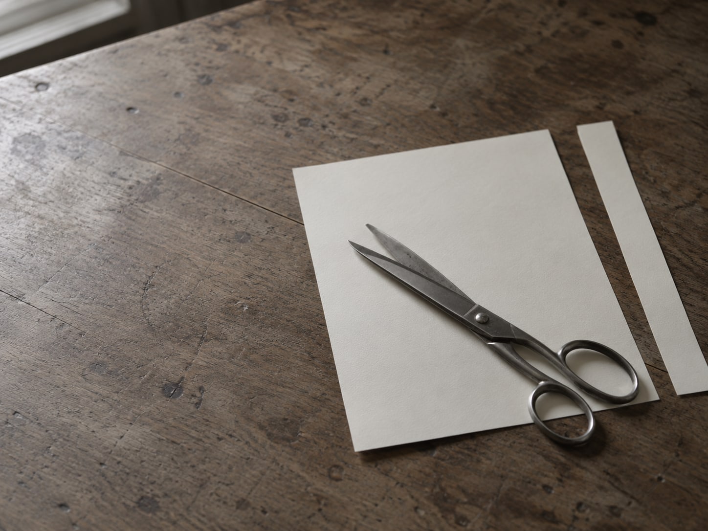 A single pair of scissors lying across a sheet of blank paper with a neat strip cut away, on a plain table.