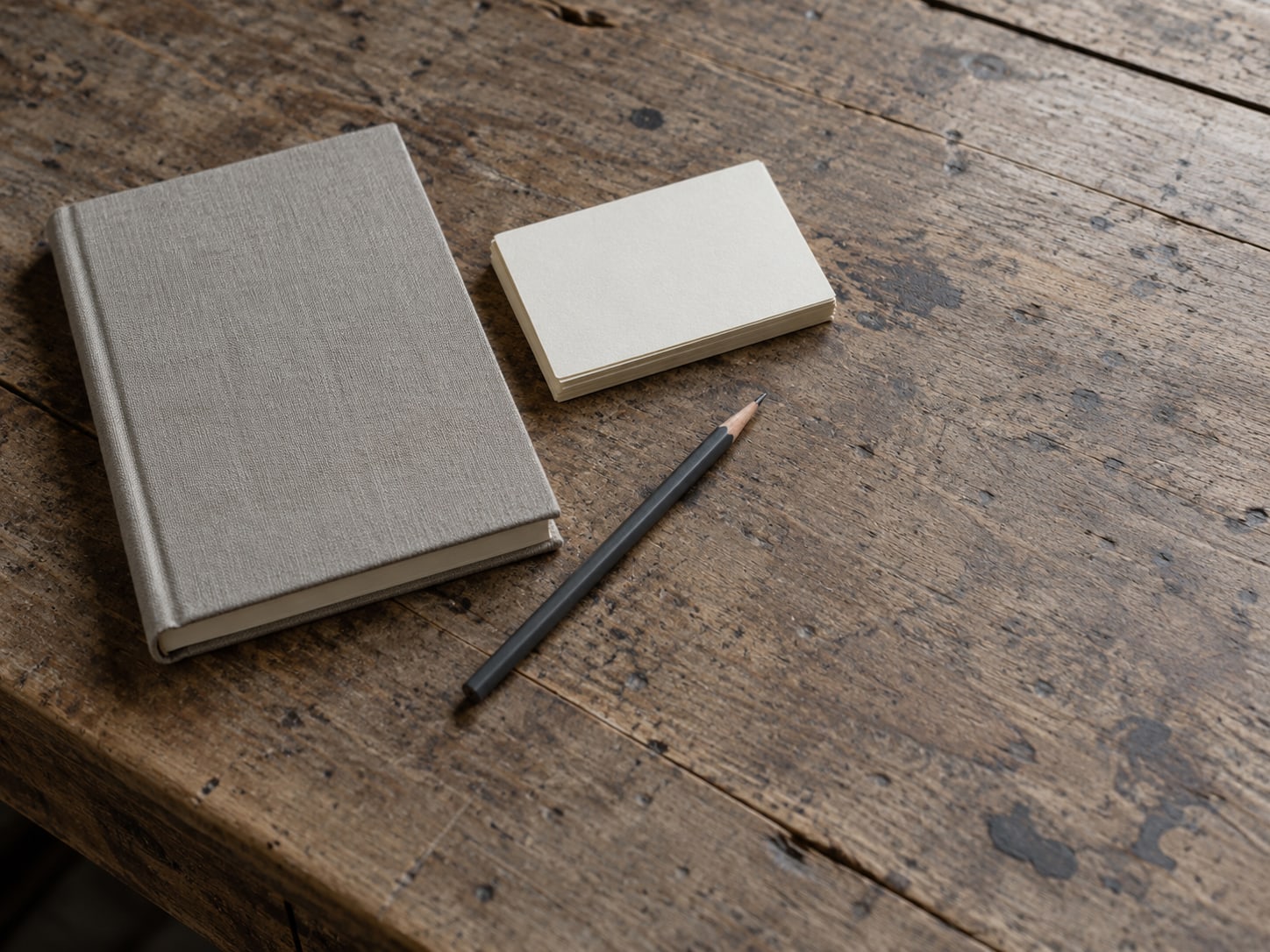 A closed notebook, a pencil and a small stack of blank index cards on a worn wooden desk.
