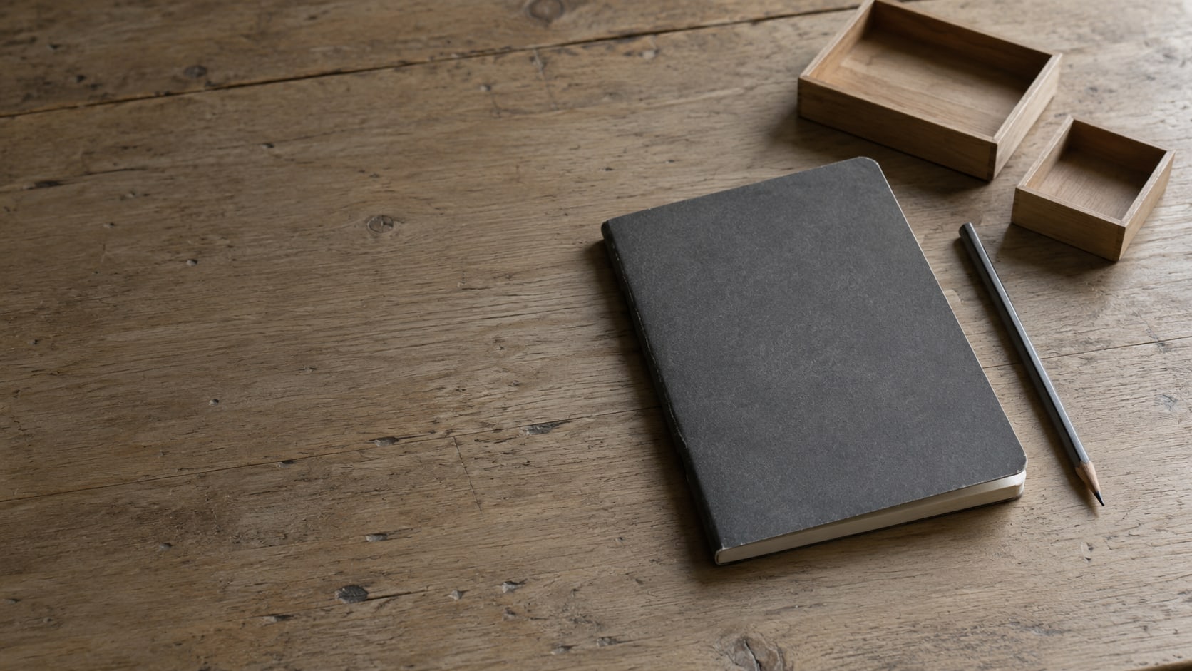 A closed notebook, a pencil and three small empty trays on a plain wooden desk.