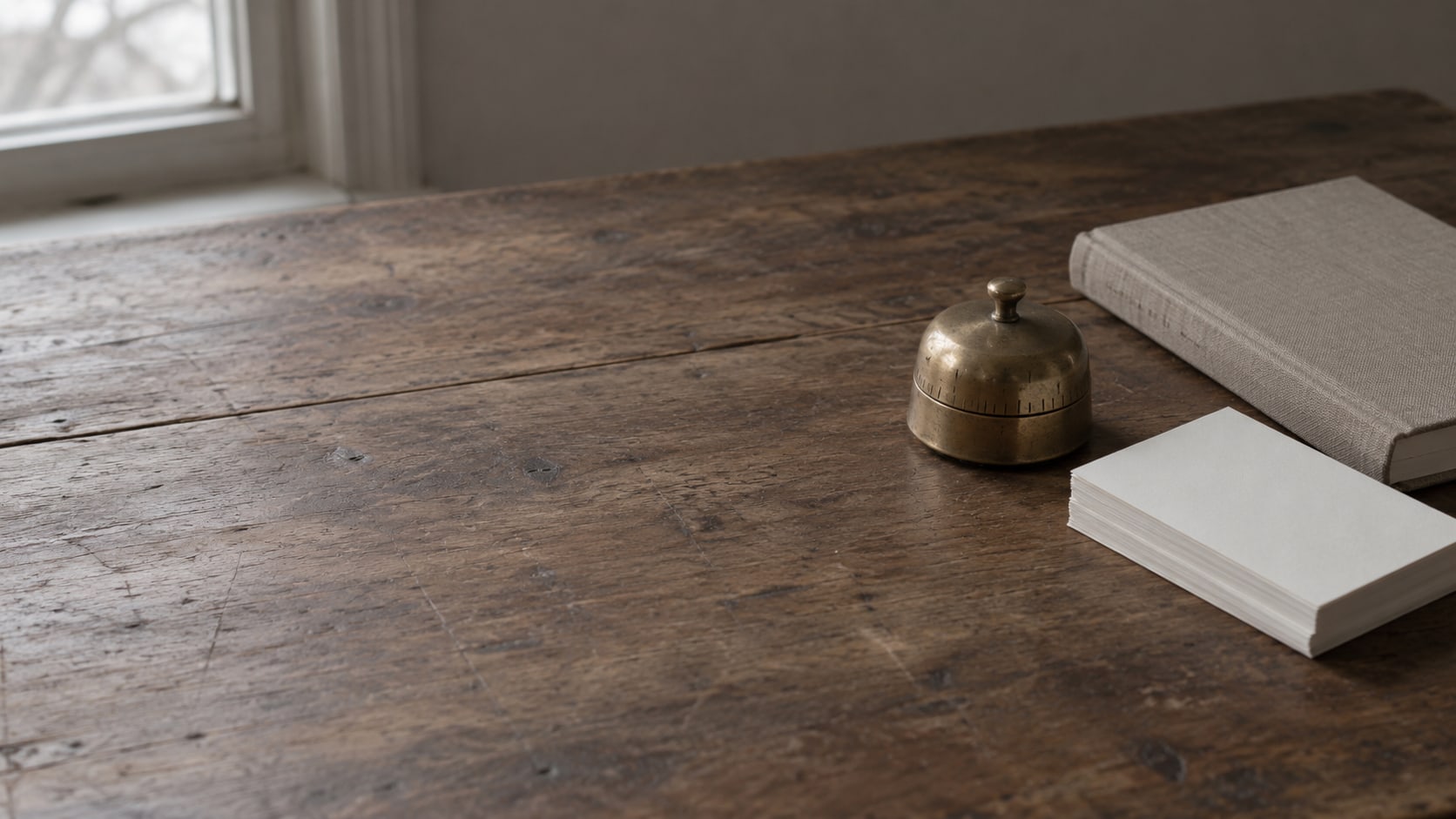 A small brass kitchen timer beside a stack of blank index cards and a closed notebook on a plain wooden desk.