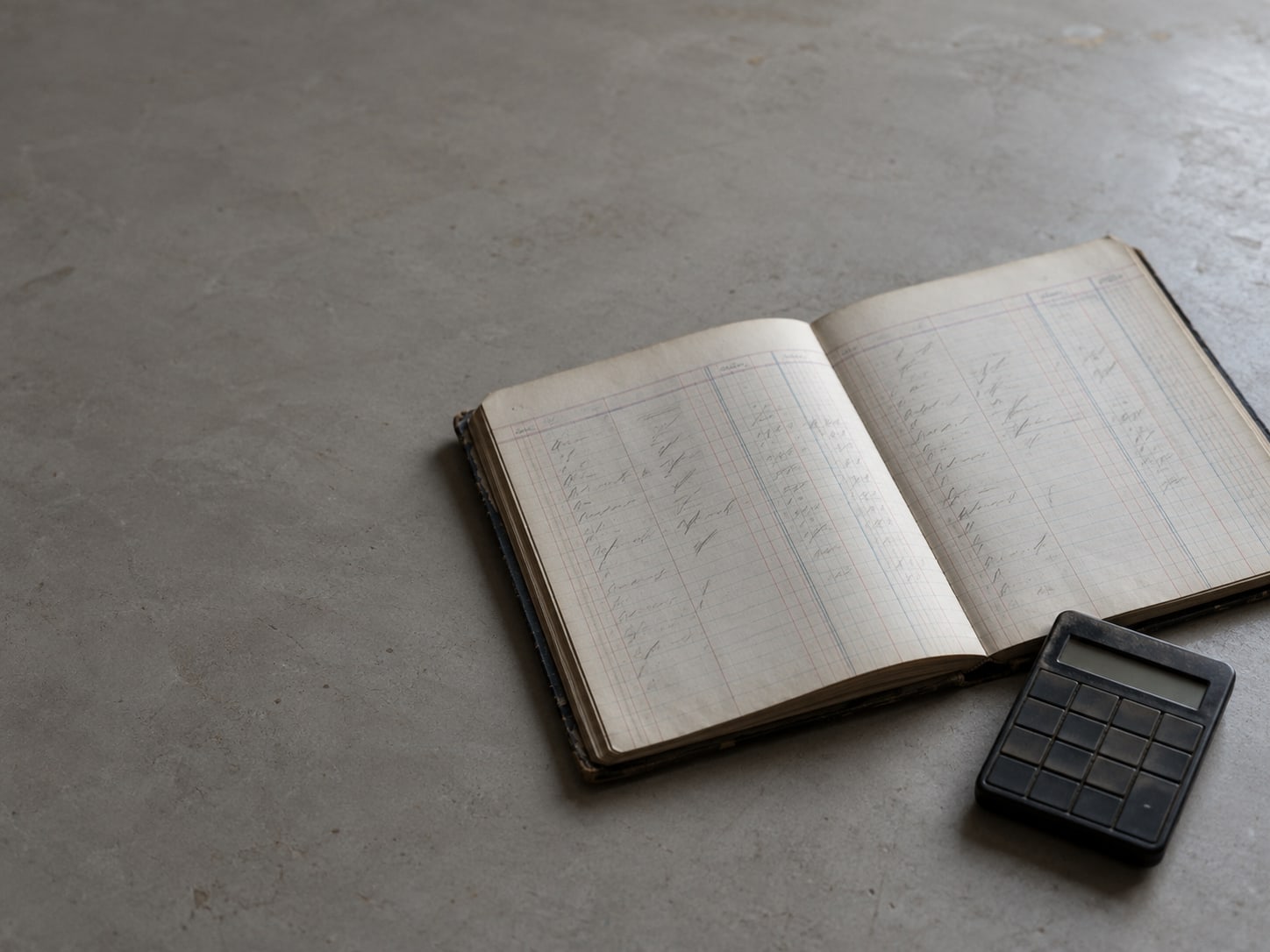A paper ledger with unreadable pencil marks in its columns beside a small flat calculator, turned off, on a plain surface.