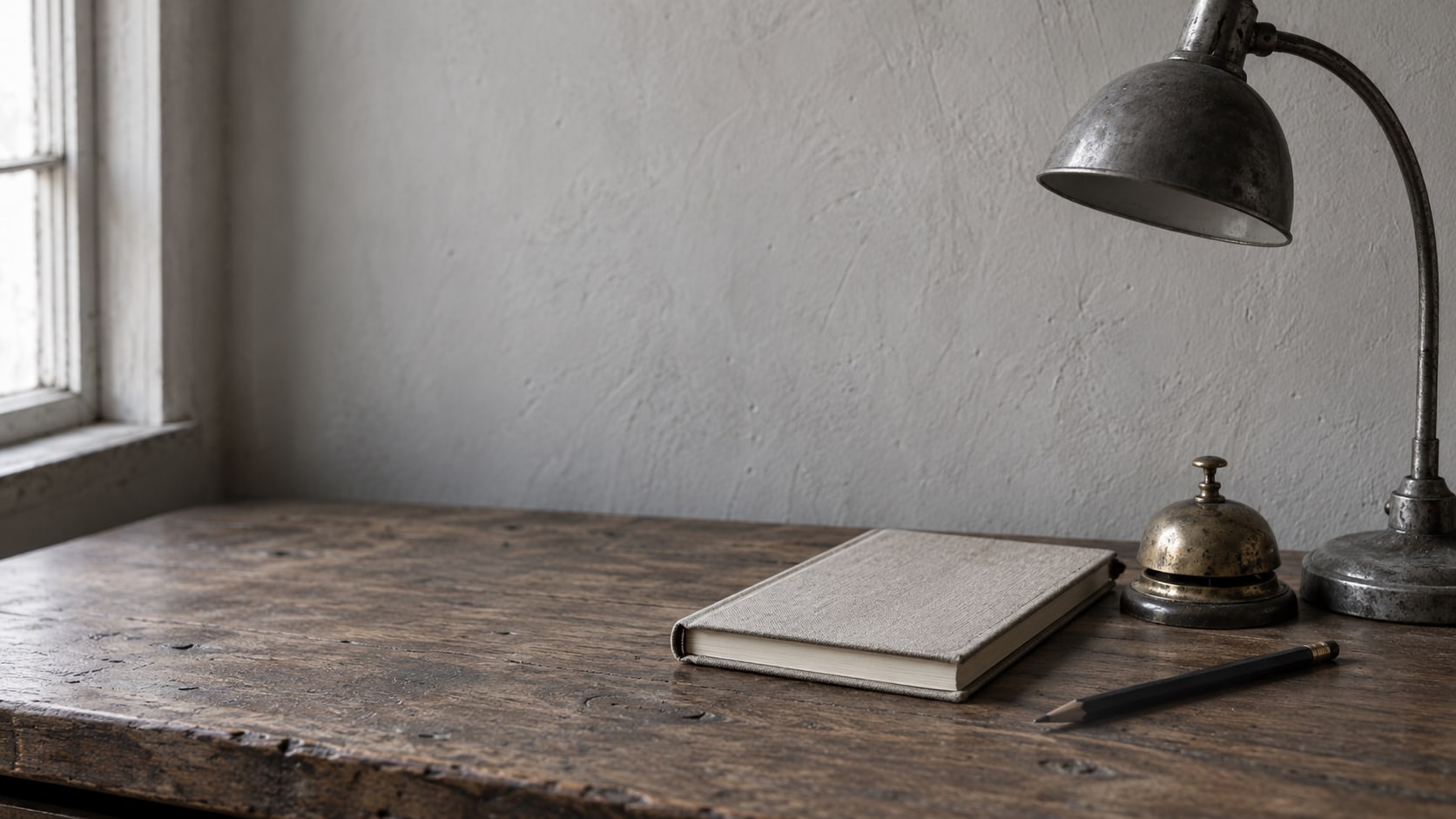 A closed notebook, a small brass bell and a pencil on a worn wooden counter beside an unlit desk lamp.
