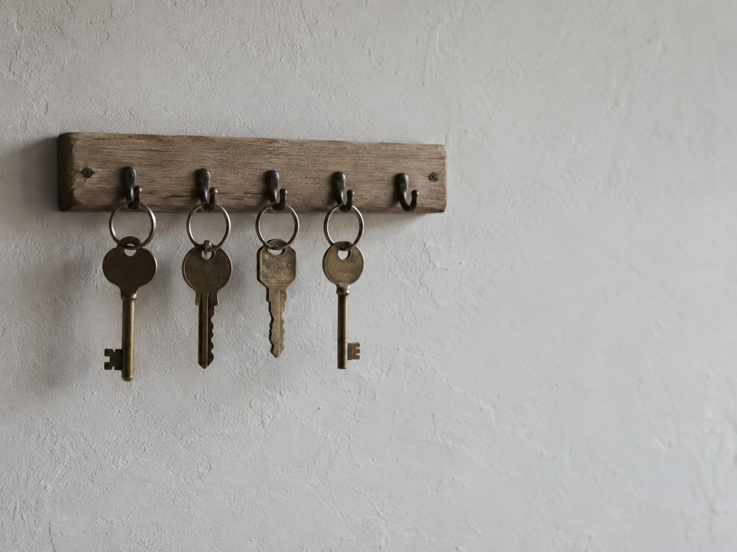 A small wooden key rack on a plain wall with four brass keys hanging from hooks and one hook left empty.