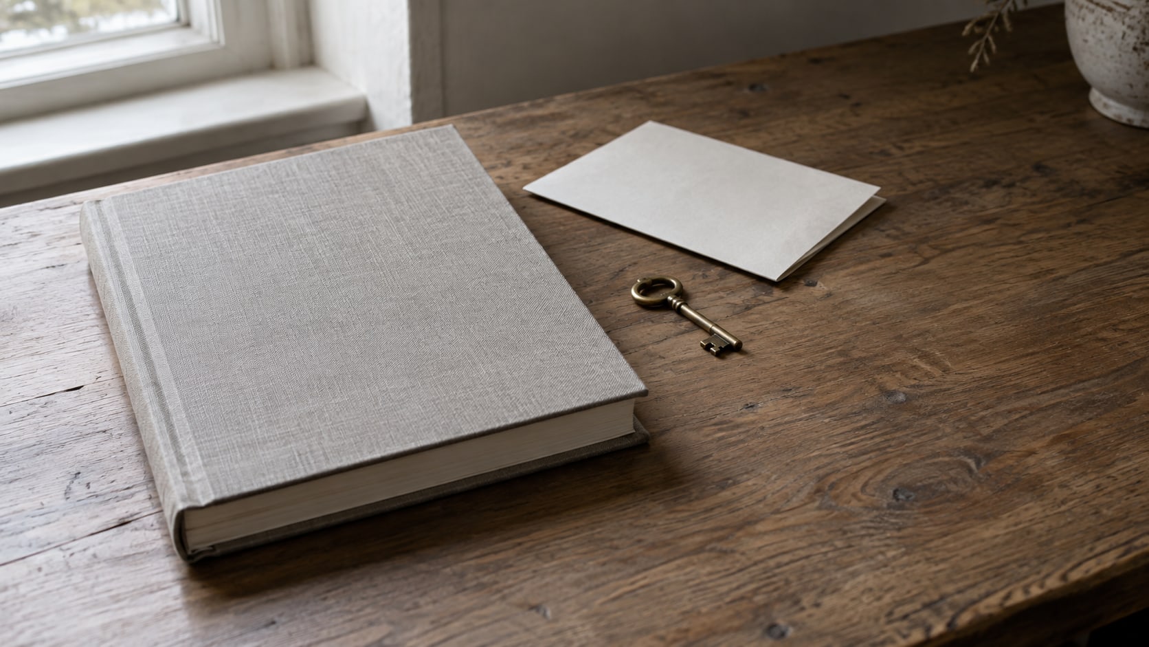 A closed, bound document on a bare wooden desk beside a brass key and a folded blank sheet of paper, lit by soft window light.