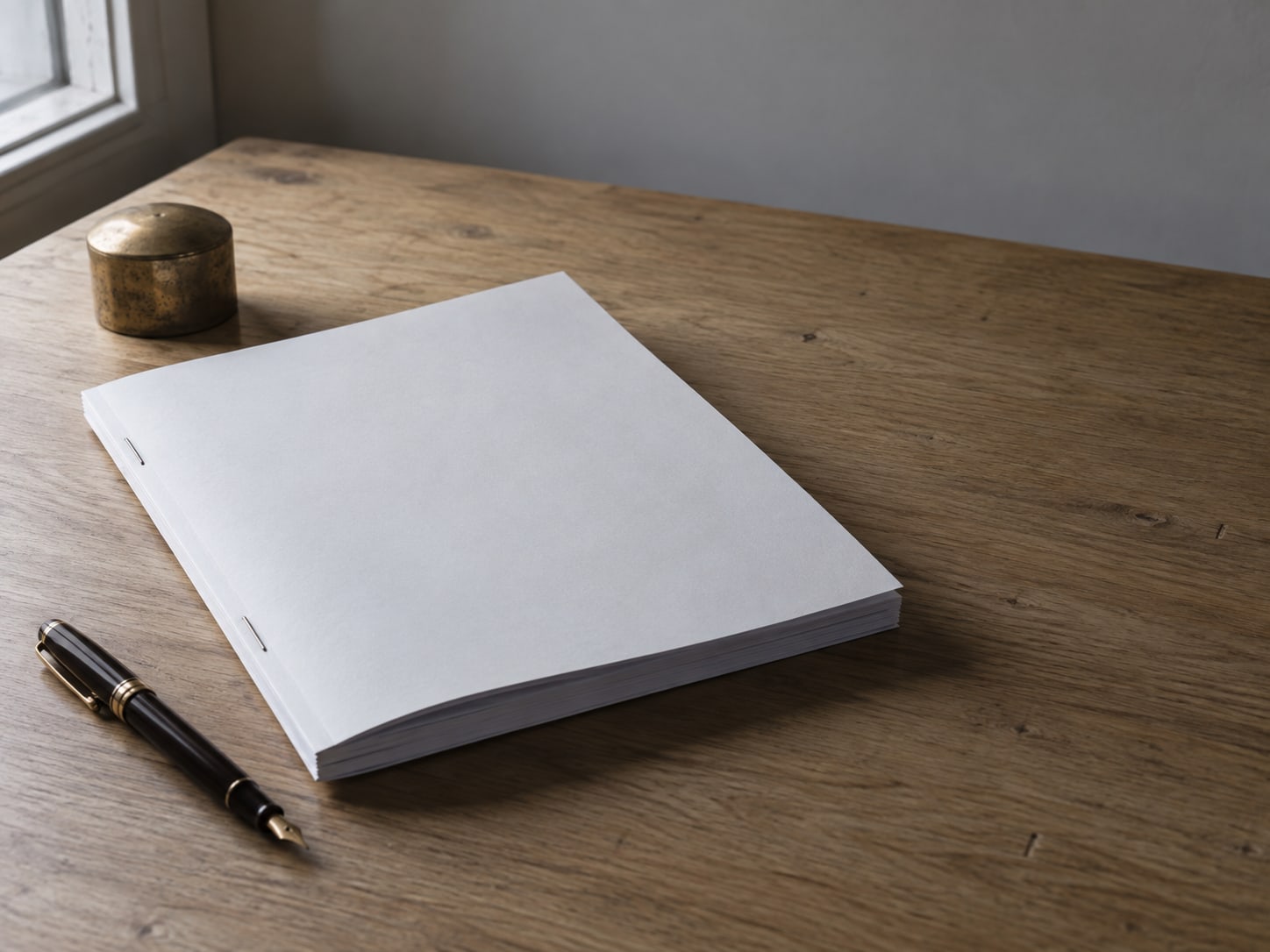 A thick stapled document lying closed on a plain oak desk beside an unused fountain pen and a small brass paperweight.