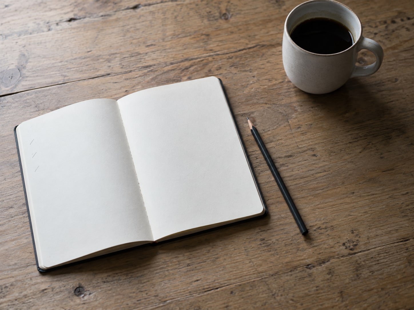 An open blank notebook on a desk with a few faint pencil tick marks in the margin, a sharpened pencil and a mug of coffee beside it.