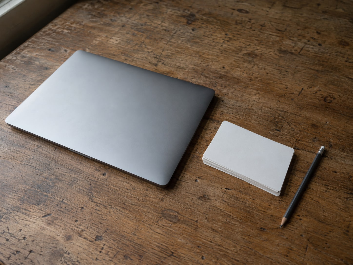 A closed laptop and a small stack of blank index cards on a wooden desk beside a pencil.
