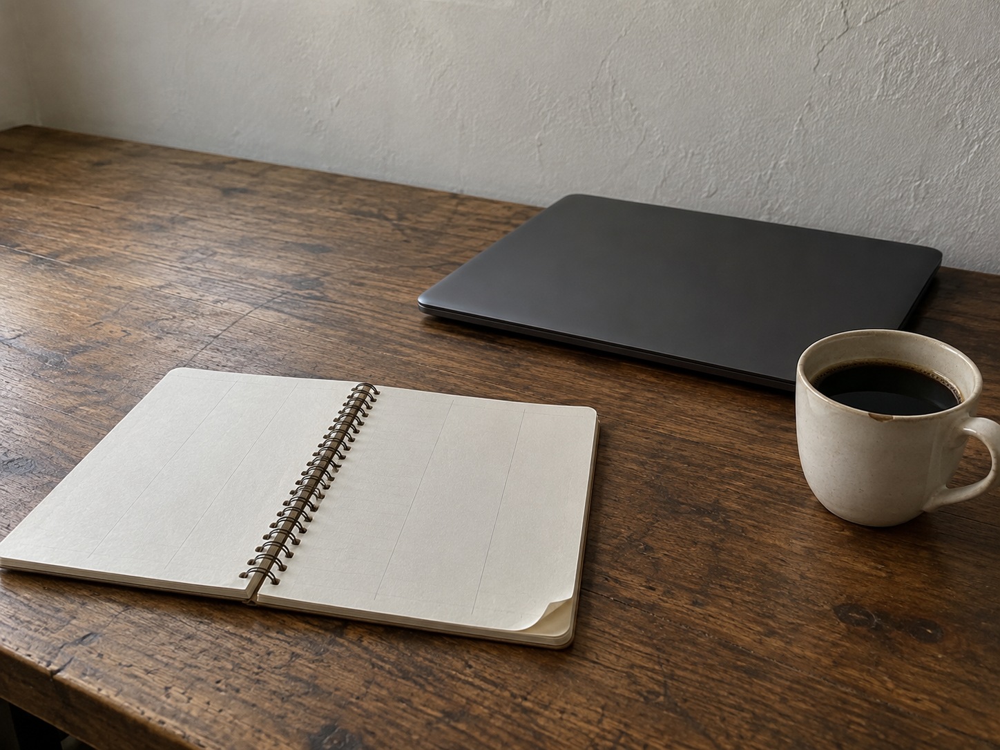 A spiral notebook open to a page of faint pencil columns beside a closed laptop and a cup of coffee on a wooden desk.