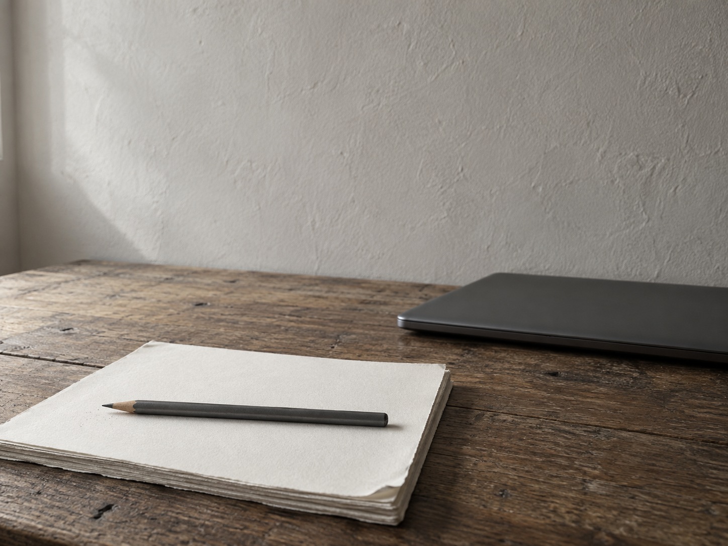 A small wooden desk with a neat stack of blank paper, a pencil laid across it and a closed laptop pushed to the side.