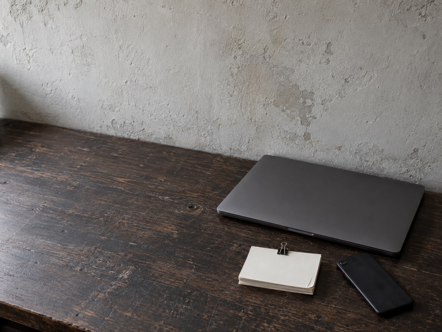 A closed laptop and a face-down phone resting on a desk beside a short stack of blank index cards held with a binder clip.