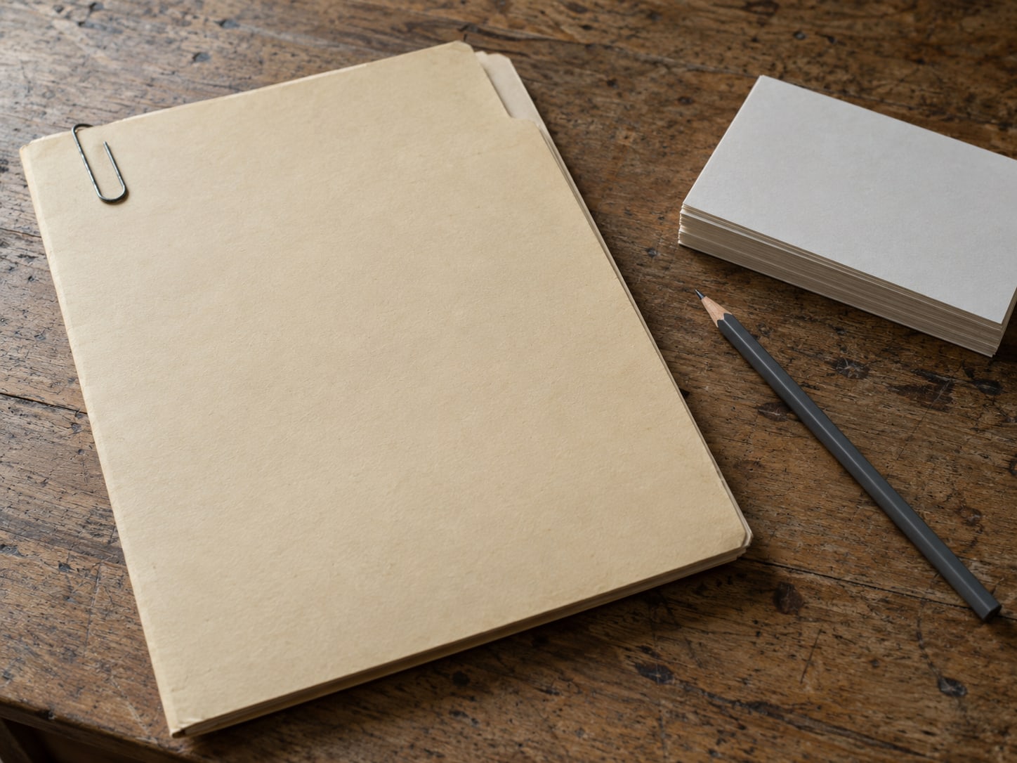 A closed manila folder with a paper clip, resting on a worn desk beside a pencil and a small stack of blank index cards.