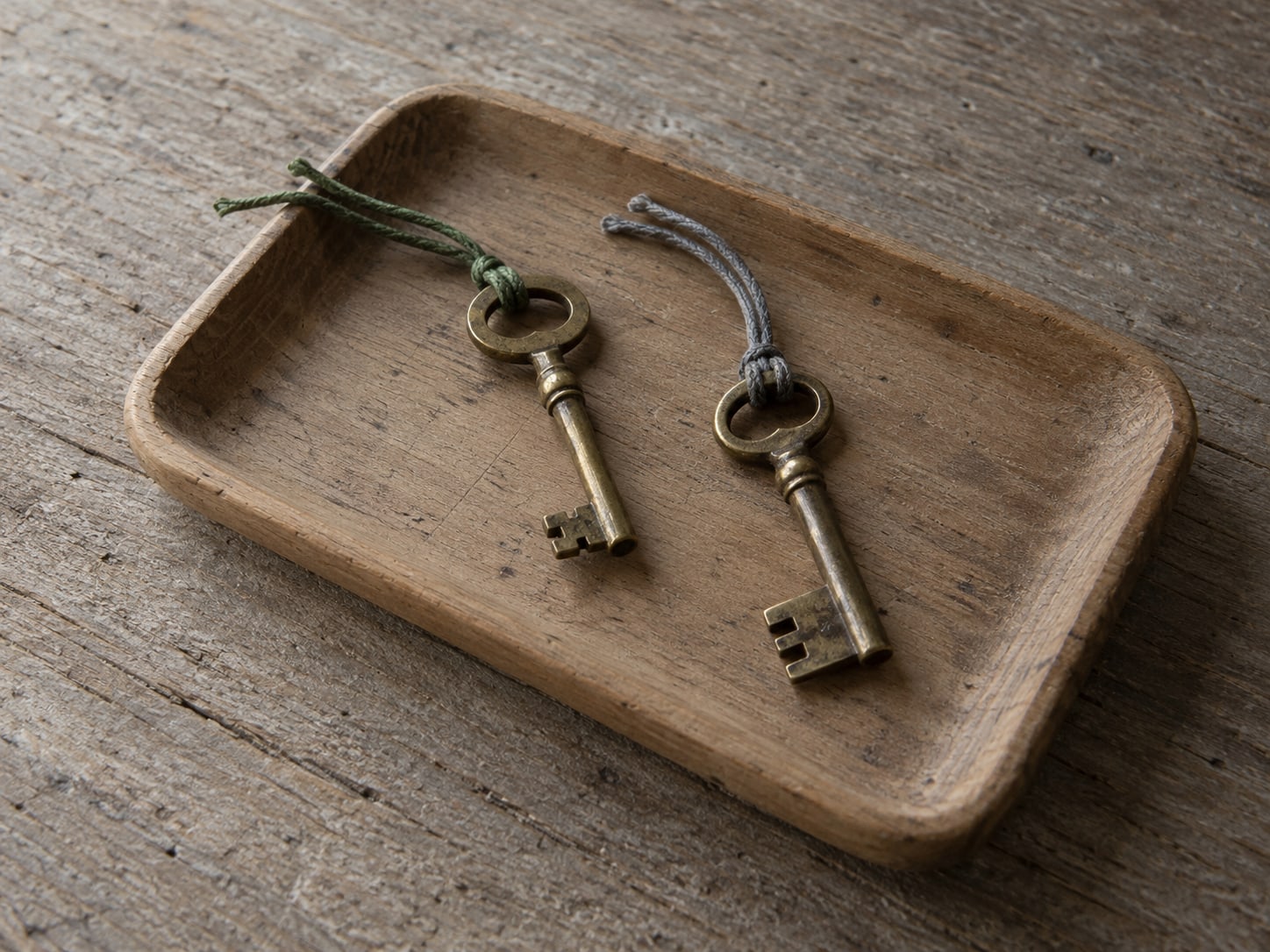 Two separate brass keys on a small wooden tray, one with a green cord and one with a gray cord.