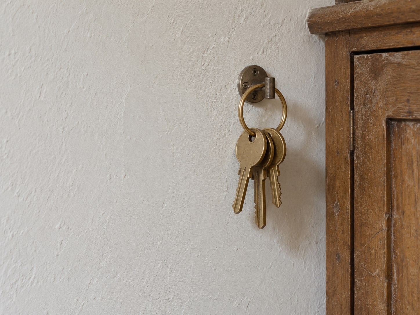 A brass key ring with three keys hanging from a wall hook beside a closed wooden cabinet.