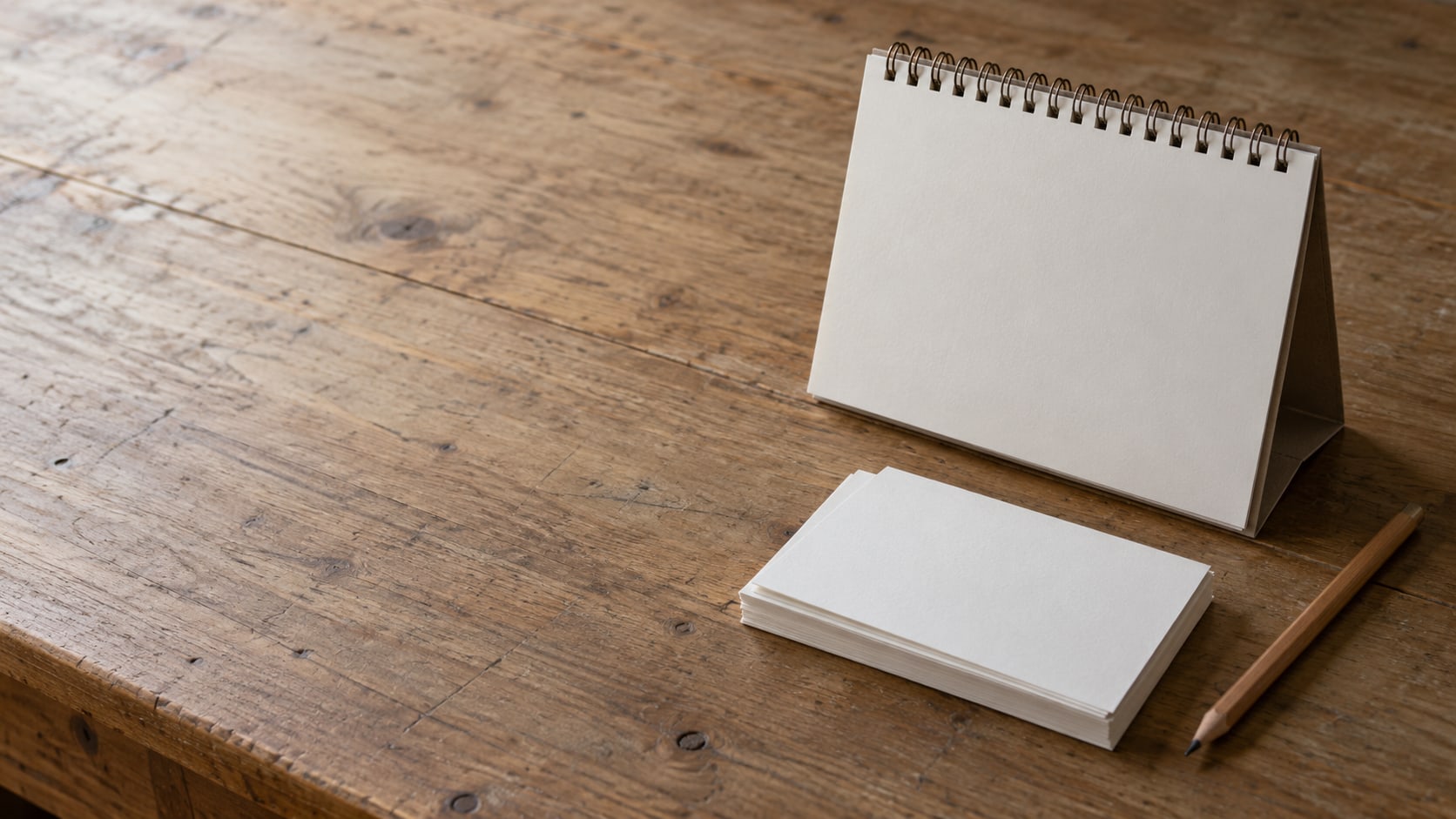 A desk calendar, a small stack of blank index cards and a sharpened pencil on a clean wooden desk.