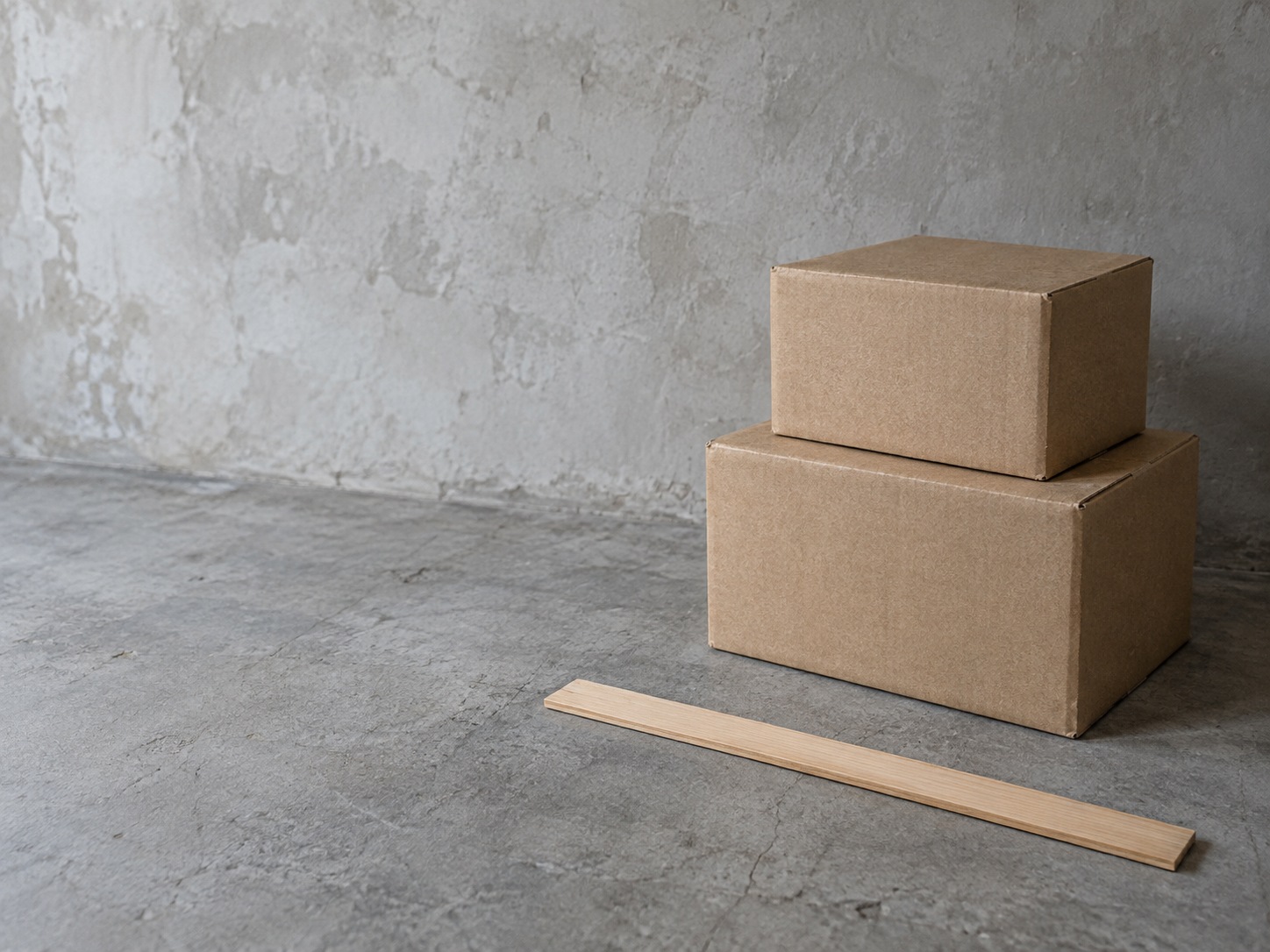 Two unmarked cardboard boxes of different sizes stacked beside a plain wooden ruler on a concrete floor.