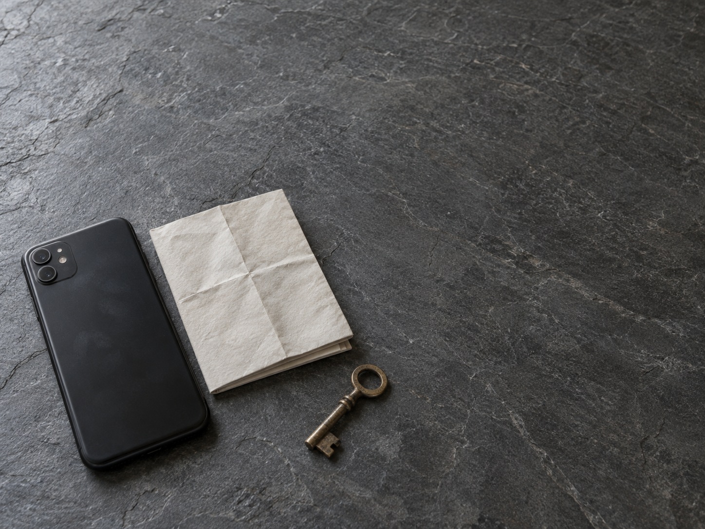 A switched-off smartphone lying face down next to a folded paper map and a brass key on a slate countertop.
