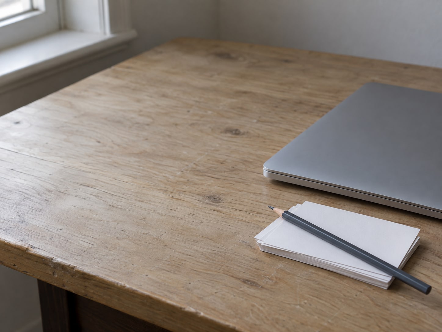 A clean wooden desk with a closed laptop, a small stack of blank index cards and a pencil laid across them.