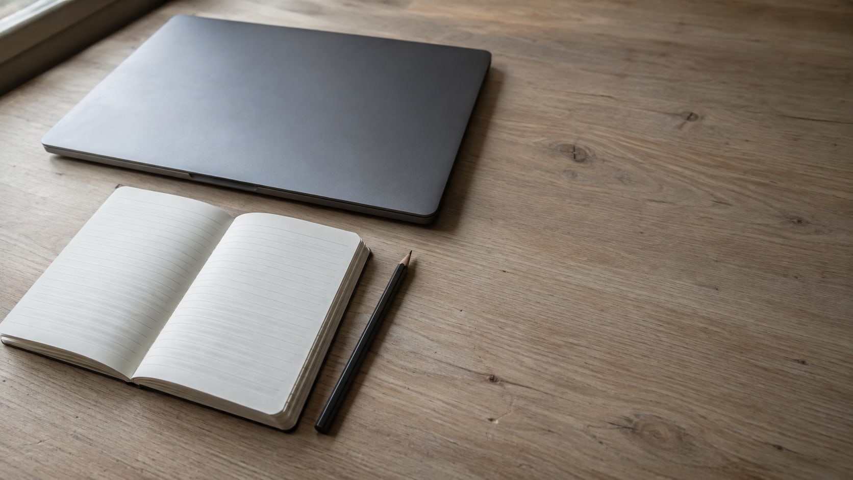 A small notepad with blank lined pages and a pencil beside a closed laptop on a tidy wooden desk.