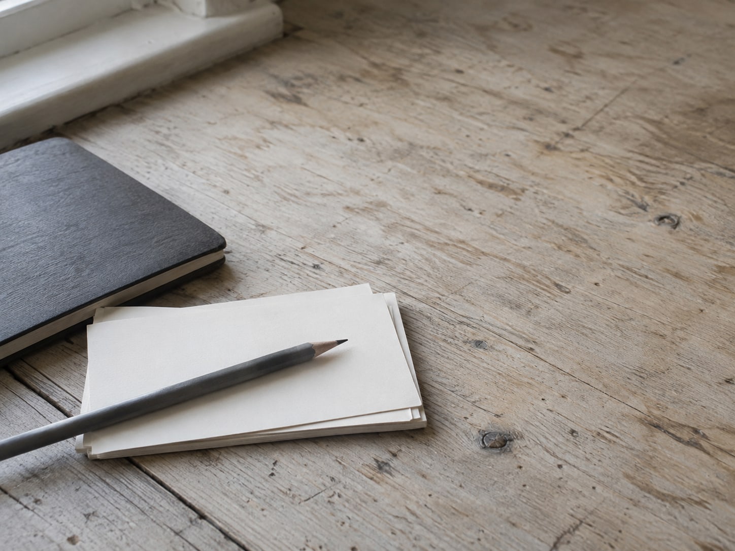 A small stack of blank index cards and a pencil resting on a wooden counter next to a closed notebook.