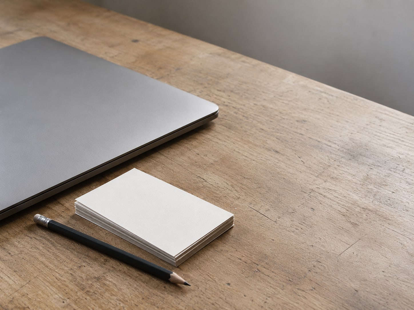A closed laptop beside a small stack of blank index cards and a pencil on a plain wooden desk.