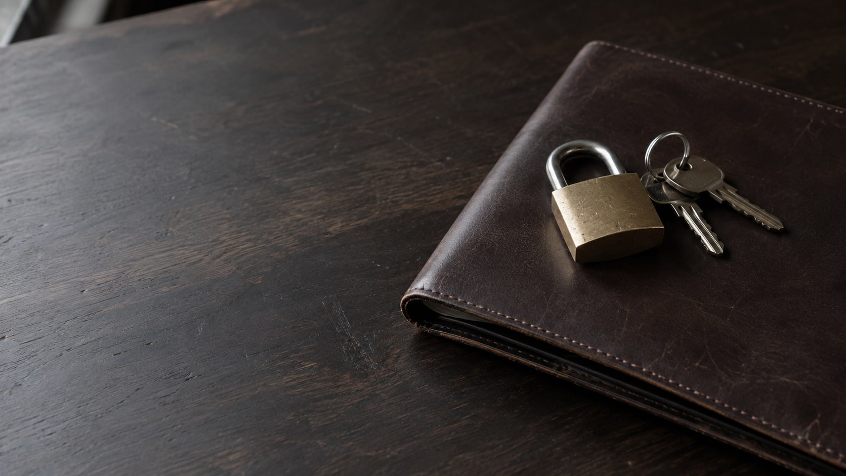 A brass padlock and a small ring of keys resting on a closed leather folder on a dark wooden desk.
