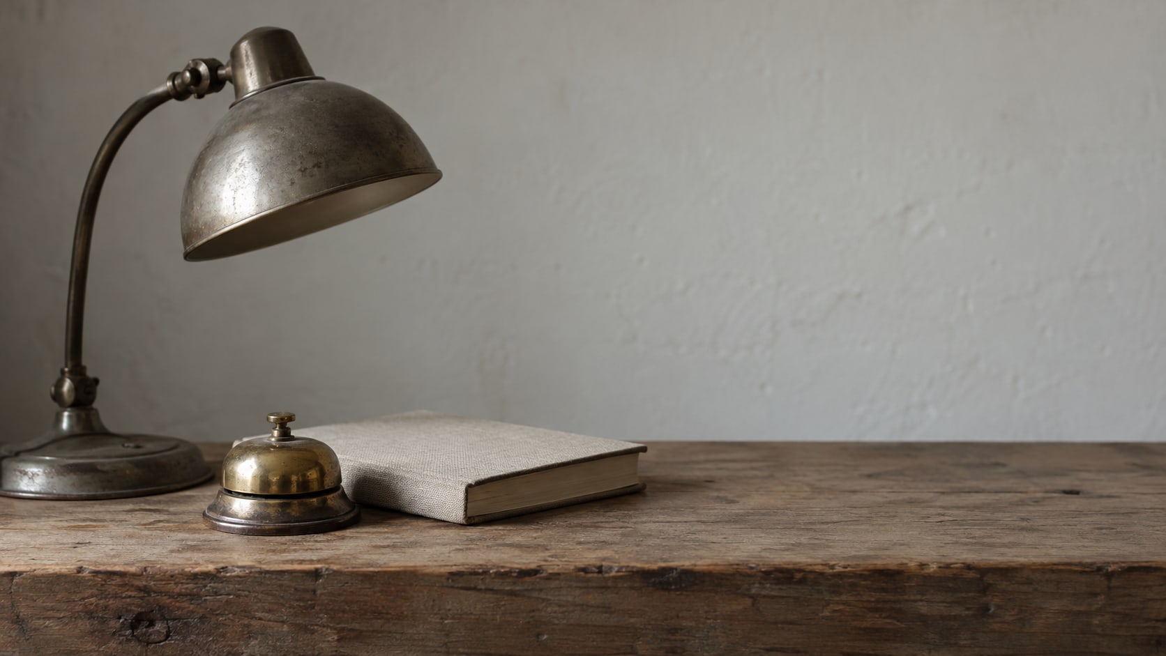 A small brass bell and a closed notebook beside an unlit desk lamp on a wooden counter.
