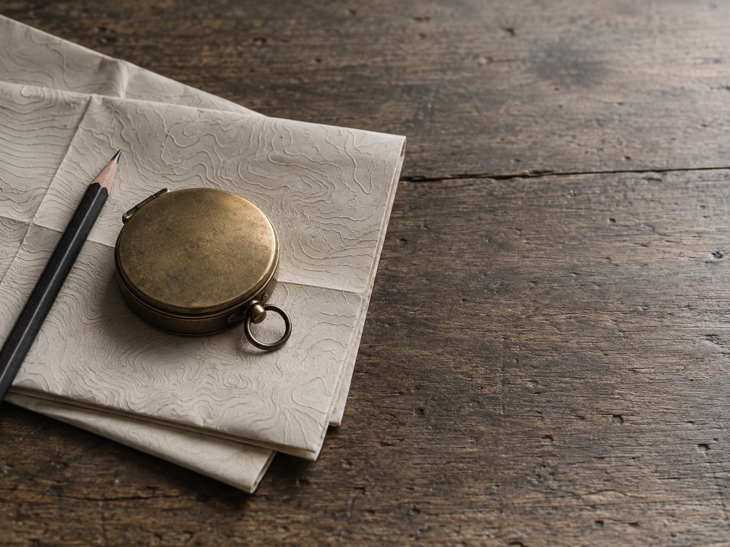 A small brass compass lying on a folded paper map beside a pencil on a worn desk.
