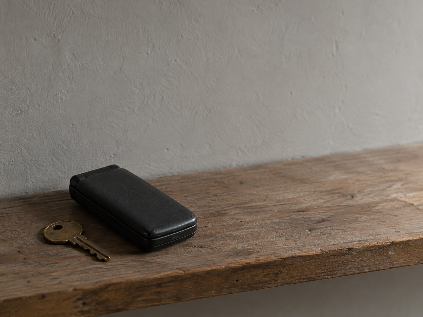 A closed flip phone resting beside a small brass mailbox key on a wooden shelf.