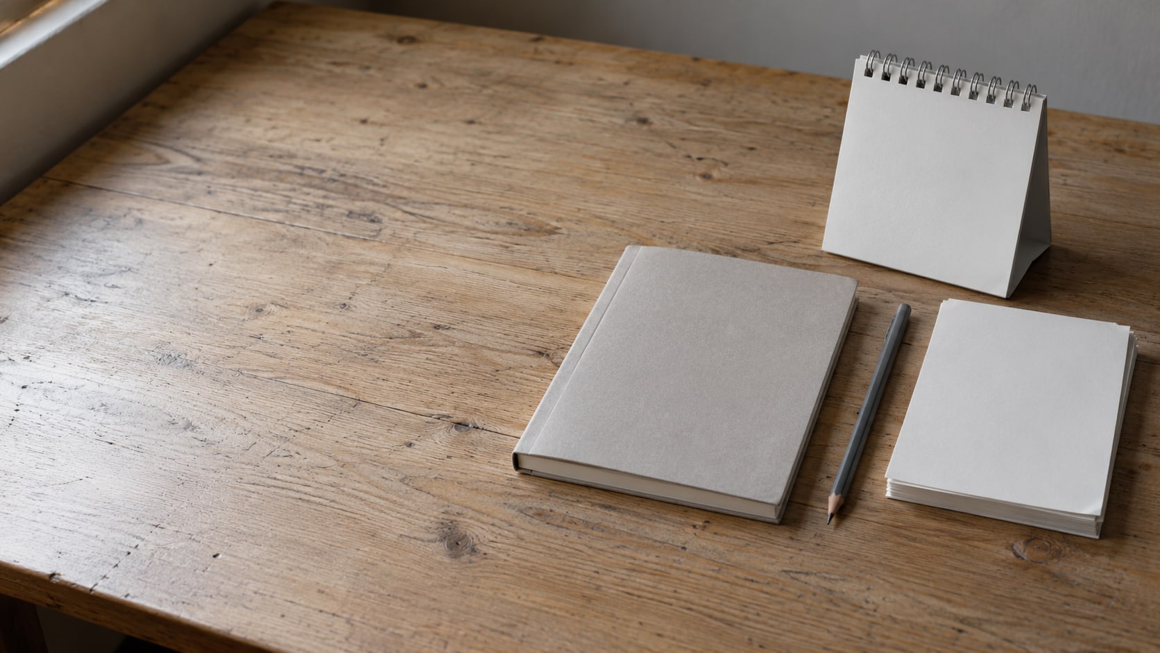 A closed notebook, a pencil and a blank paper desk calendar beside a small stack of plain paper on a wooden desk.