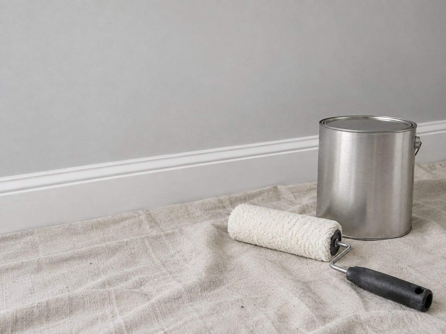 A clean paint roller and a closed paint can on a drop cloth beside a neatly finished baseboard.