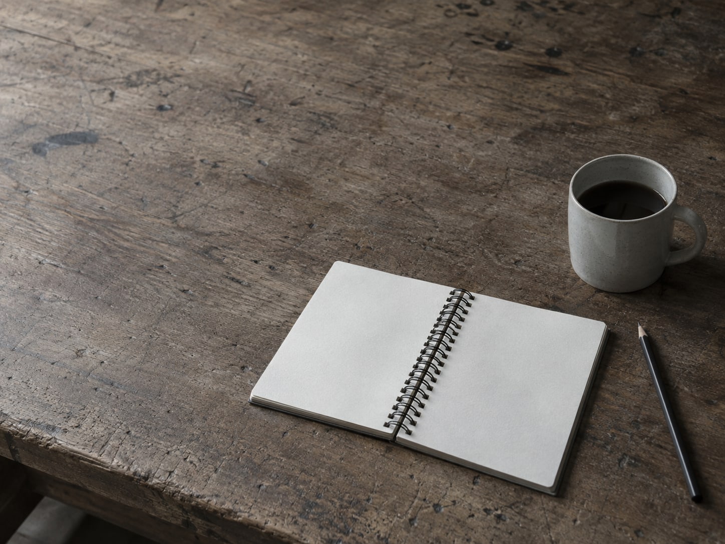 Un cuaderno de espiral abierto en páginas en blanco junto a un lápiz y una taza de café sobre un banco de trabajo de madera desgastada.