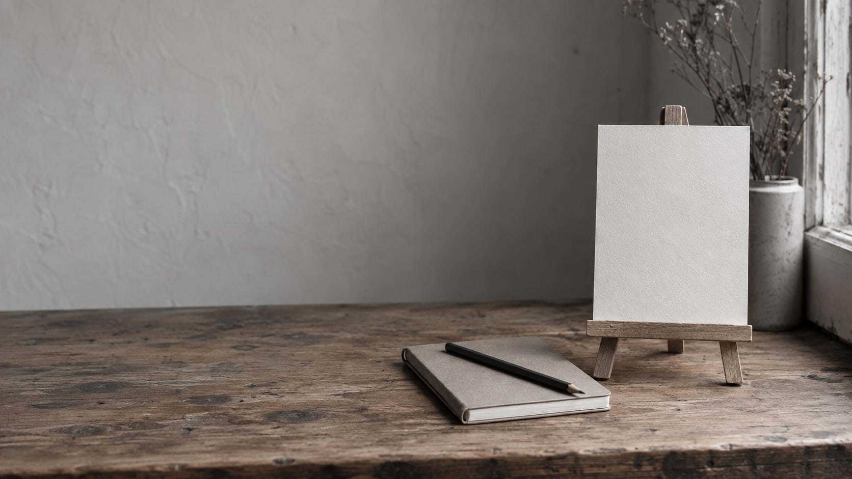 Un letrero de papel en blanco sobre un pequeño caballete junto a un cuaderno cerrado y un lápiz sobre un mostrador de tienda de madera.