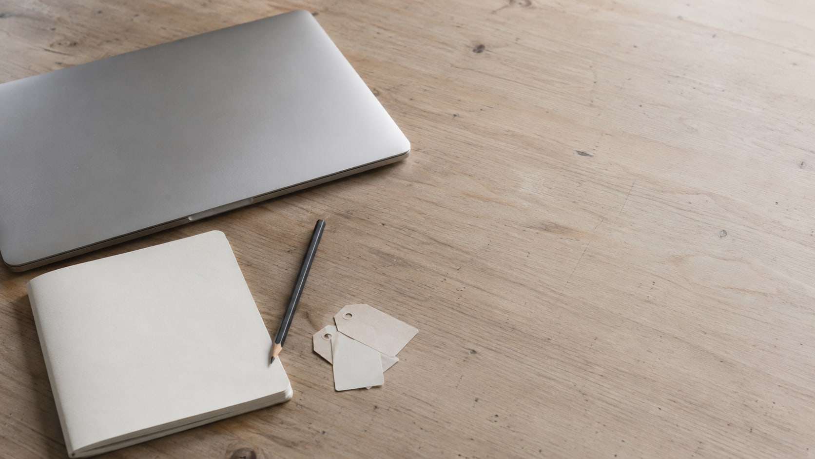 A closed laptop and a pencil beside a plain notebook and a few small paper tags on a light wooden desk.