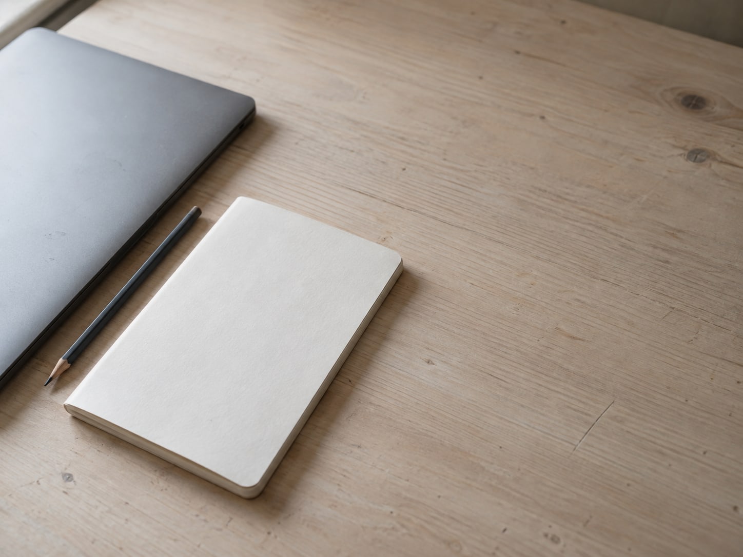 A closed laptop and a pencil lying beside a plain notebook on a light wooden desk.