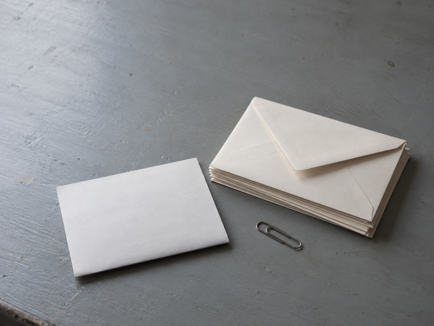 A small stack of blank unopened envelopes beside a folded sheet of paper and a paperclip on a gray desk.