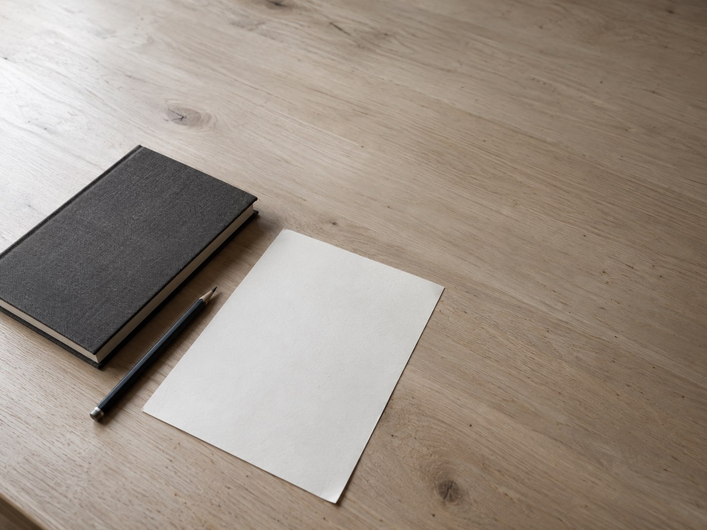 A single sheet of blank paper beside a pencil and a closed notebook on a tidy desk.