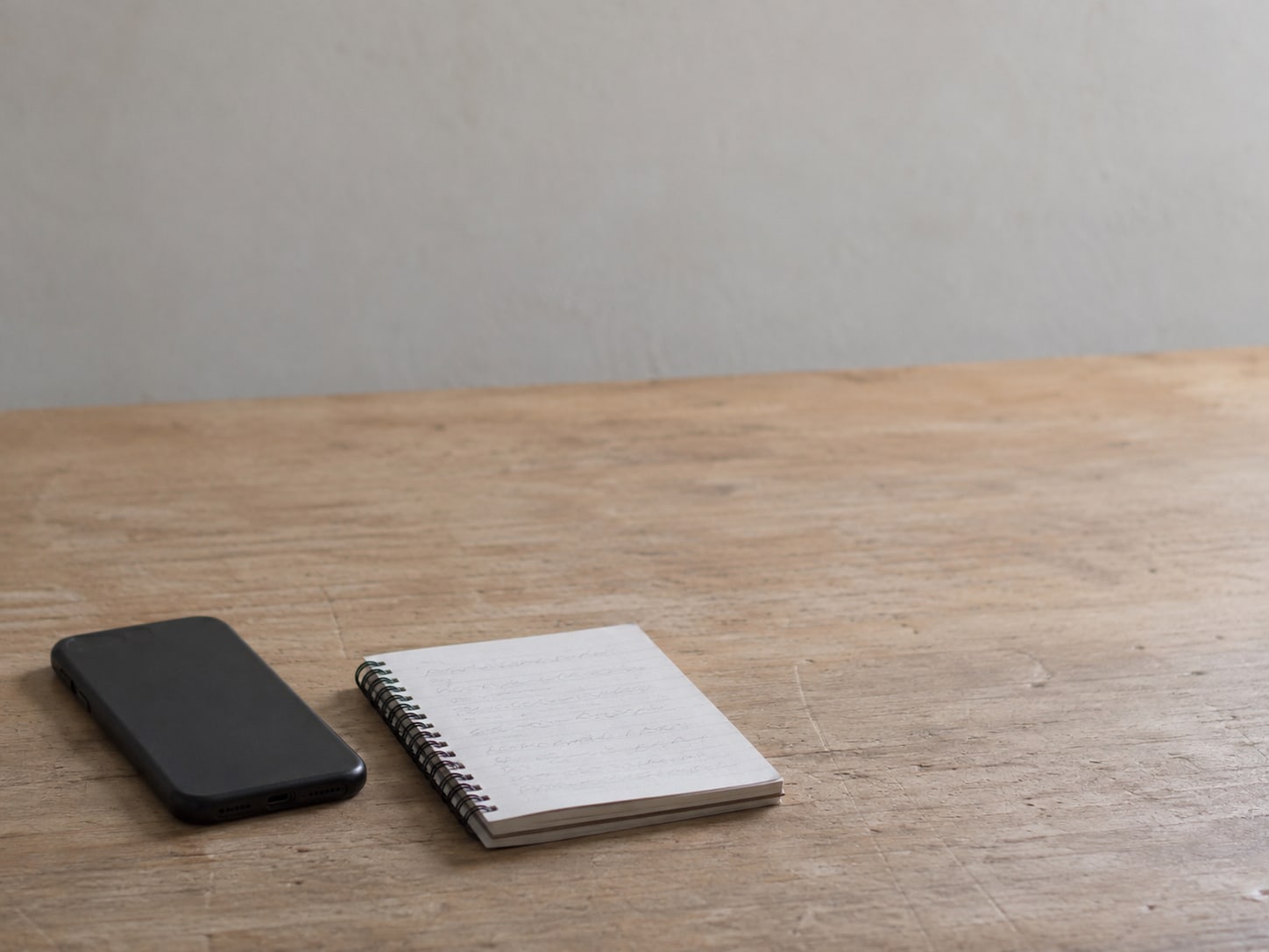 A face-down phone and a small spiral notebook with unreadable pencil marks resting on a wooden kitchen table.