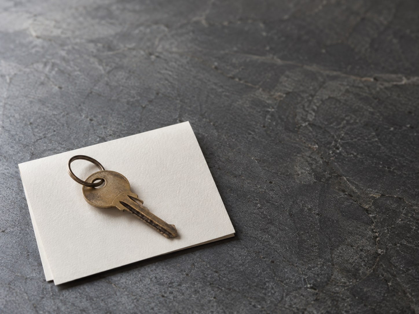 A single brass key on a plain ring resting on a folded blank card on a slate countertop.