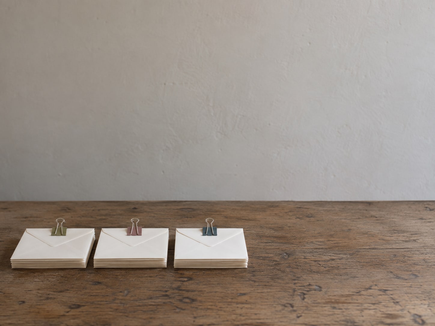 Three small stacks of blank envelopes laid out in a row on a desk, each held with a different colored binder clip.