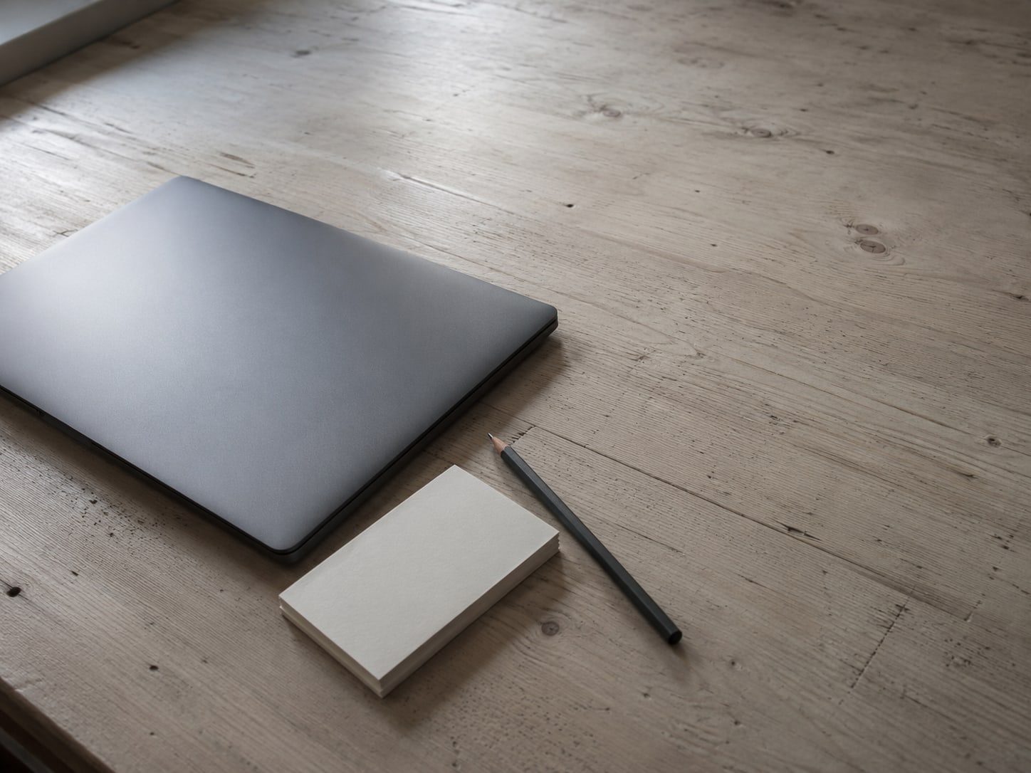 A closed laptop beside a small stack of blank index cards and a pencil on a pale wooden desk.