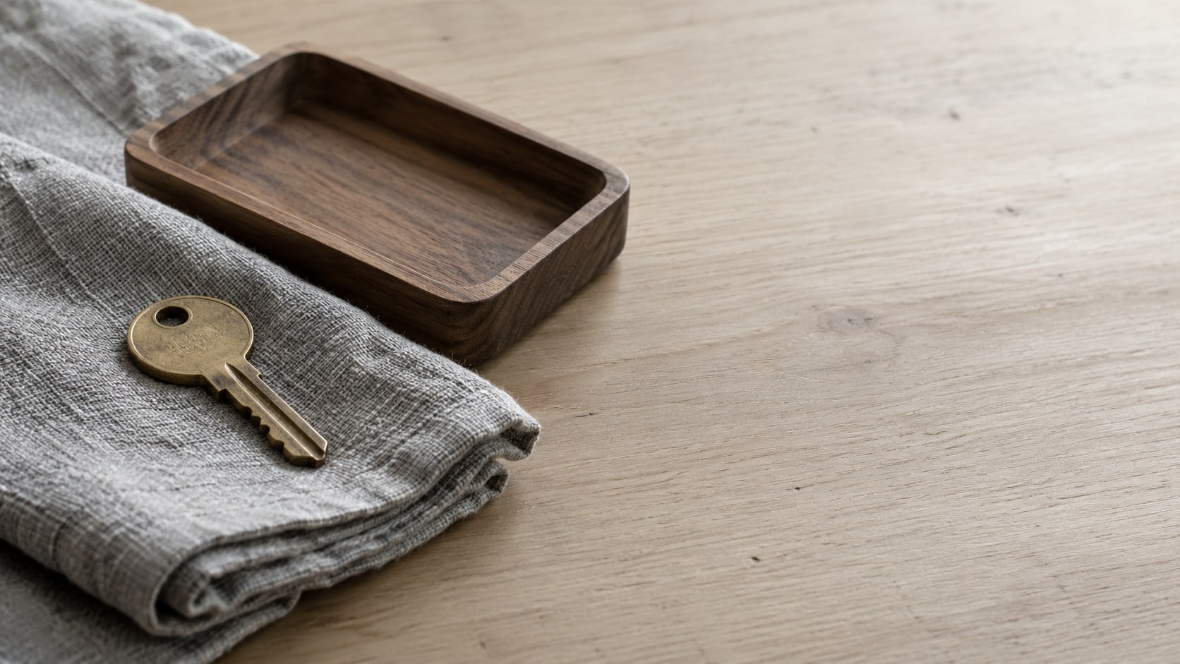 A single brass key lying on a folded gray cloth beside a small empty wooden tray on a plain table.