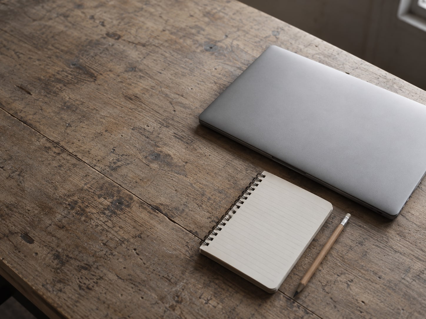 A closed laptop beside a small notepad with blank lines and a pencil on a plain wooden desk.
