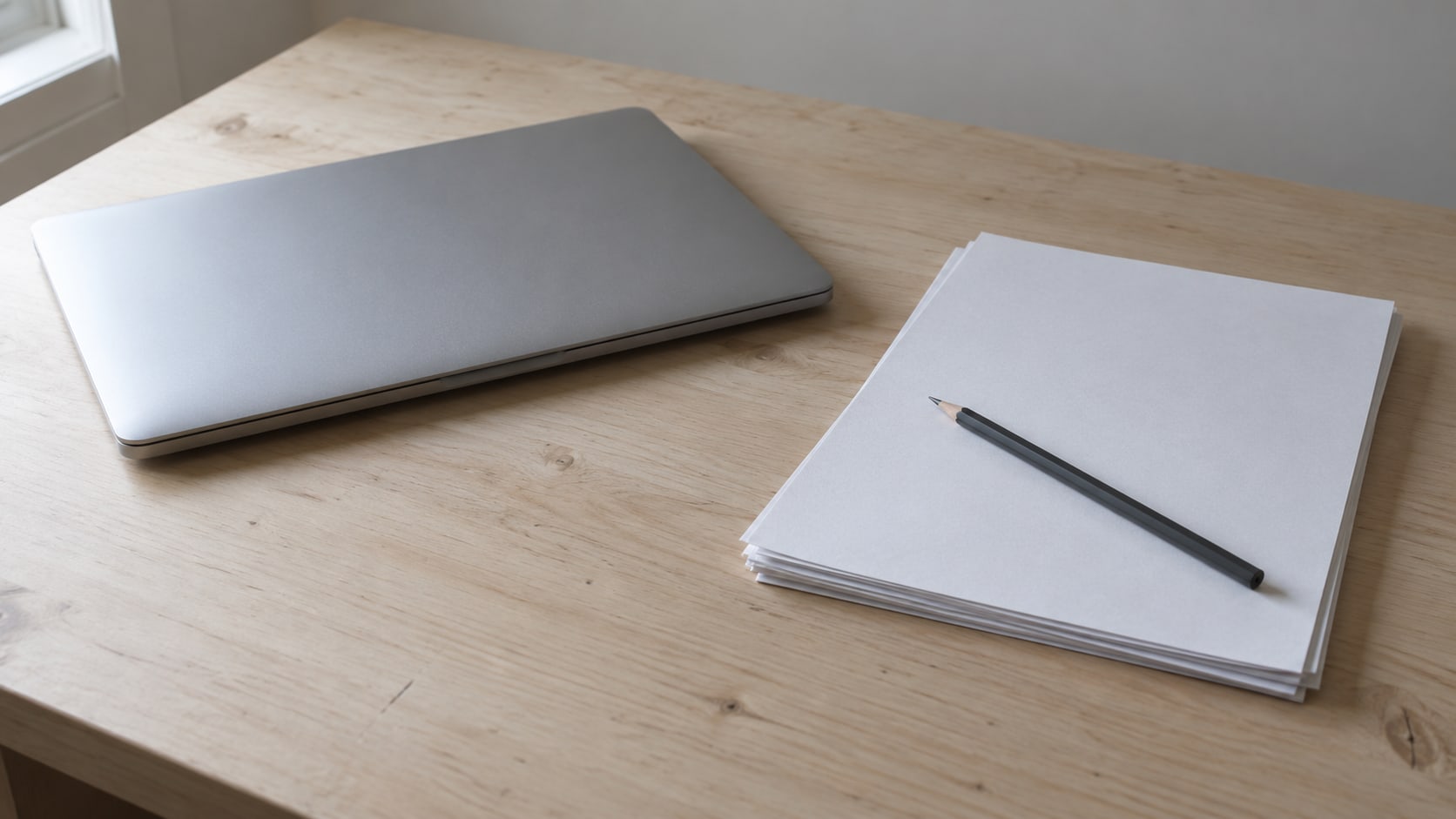A closed laptop beside a stack of blank pages and a pencil on a pale wooden desk.