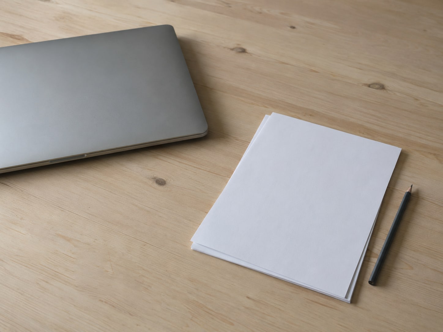 A closed laptop beside a stack of blank paper and a pencil on a pale wooden desk.