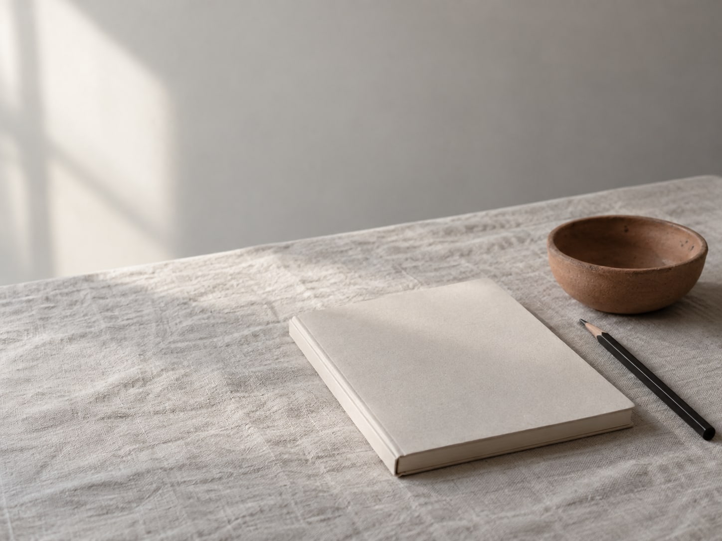 A closed notebook and a pencil resting beside a small terracotta bowl on a plain linen cloth.